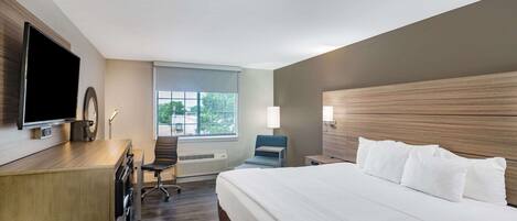 A hotel room with a large bed, a desk with a flat-screen TV, a chair, and a window with a view of trees.