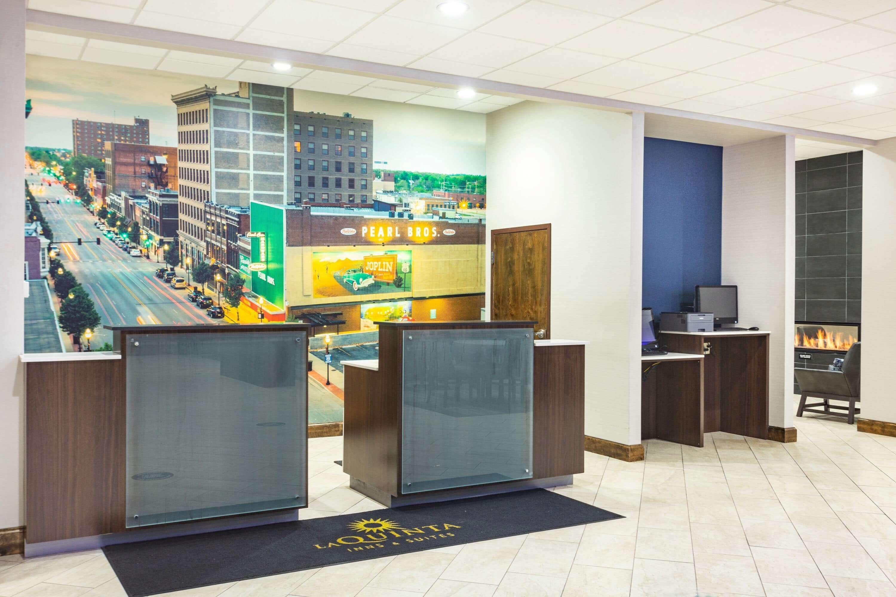 Een hotel receptie met een groot stadsgezicht schilderij, twee receptiebalies, een open haard en een zwarte deurmat met de naam van het hotel.