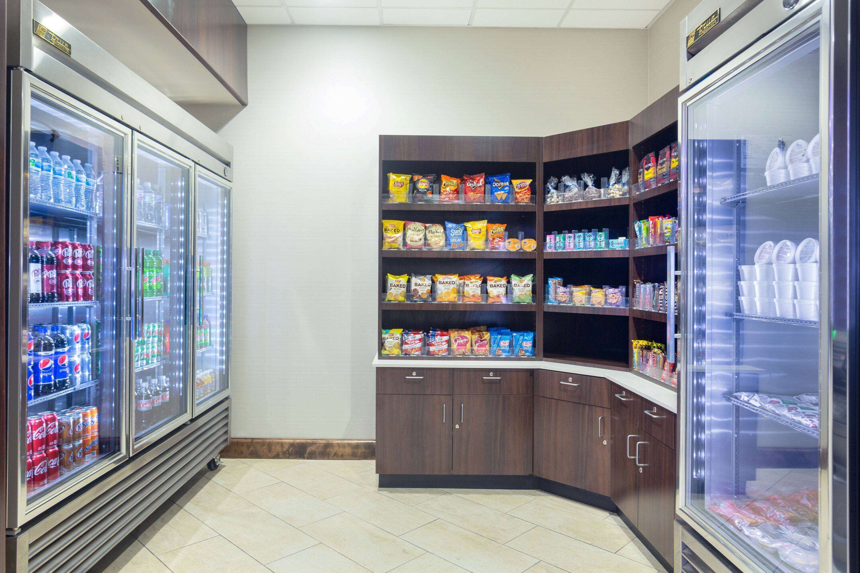 A corner of a store with refrigerated beverage dispensers, snack shelves, and a refrigerated section with cups.