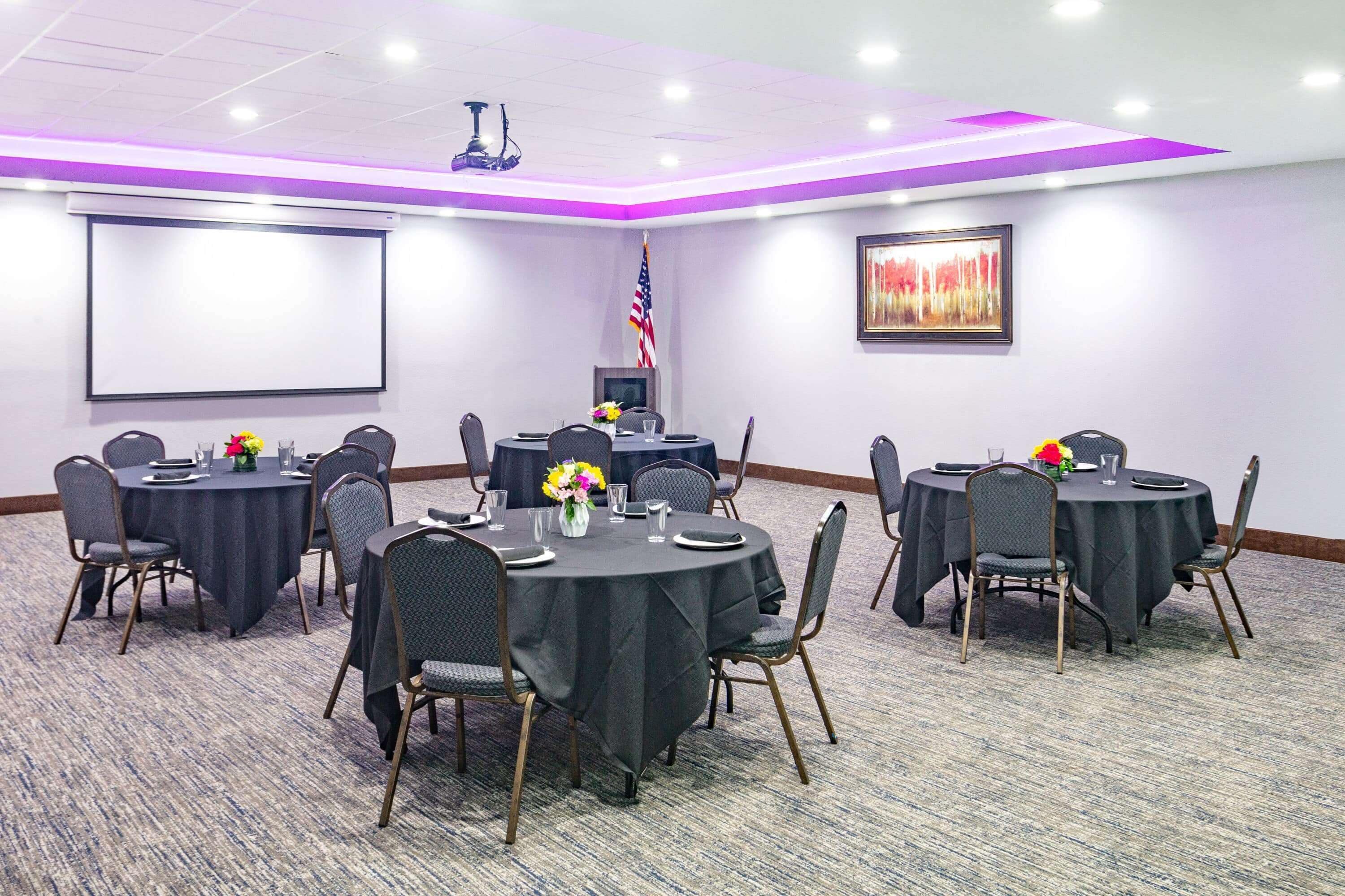 A conference room with round tables, chairs, a projector screen, an American flag, and a framed artwork on the wall.