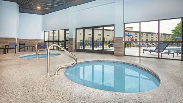 Una piscina interior con paredes de vidrio transparente, un área de descanso con sillas y vistas a un edificio cercano.