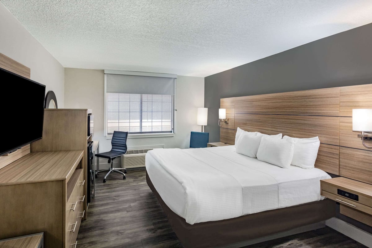 A hotel room with a large bed, a desk with a flat-screen TV, a chair, a window with blinds, and a wall-mounted lamp.