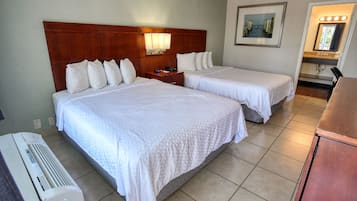 Habitación de hotel con dos camas, cabecera de madera, mesita de noche, lámpara, un cuadro y baño con tocador y espejo.