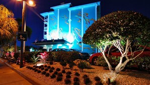 Un hotel con fachada azul, un cartel que indica 'MOTELS' con una flecha, y un jardín bien cuidado con arbustos y un árbol.