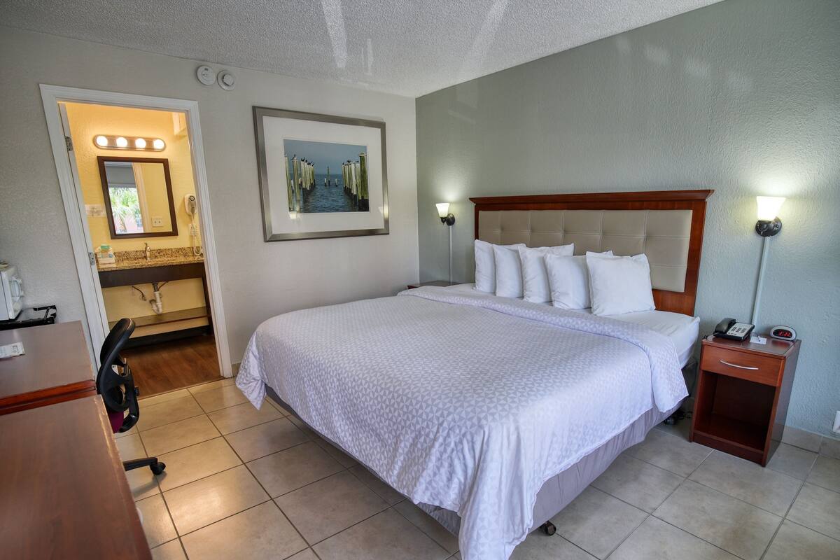 Een hotelkamer met een groot bed, een nachtkastje met een lamp, een wanddecoratie in een lijst en een badkamer die door een geopende deur zichtbaar is.