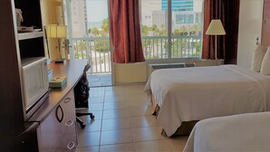 Habitación de hotel con dos camas, un escritorio con lámpara, televisión y vistas a la ciudad a través de amplias ventanas.