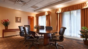 A conference room with a large table surrounded by chairs, large windows with curtains, and a potted plant on the side.