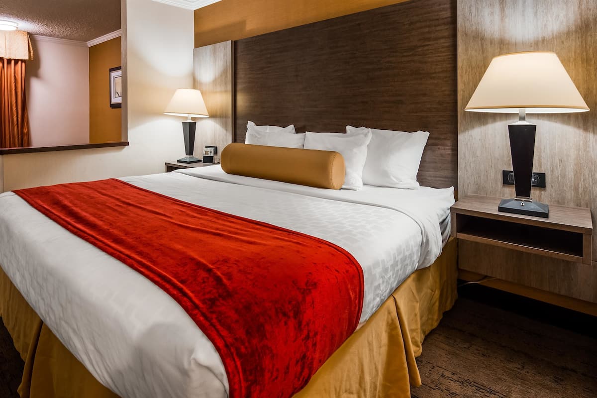 A hotel room with a large bed, two bedside tables, a nightstand, a red throw blanket, and two lamps.