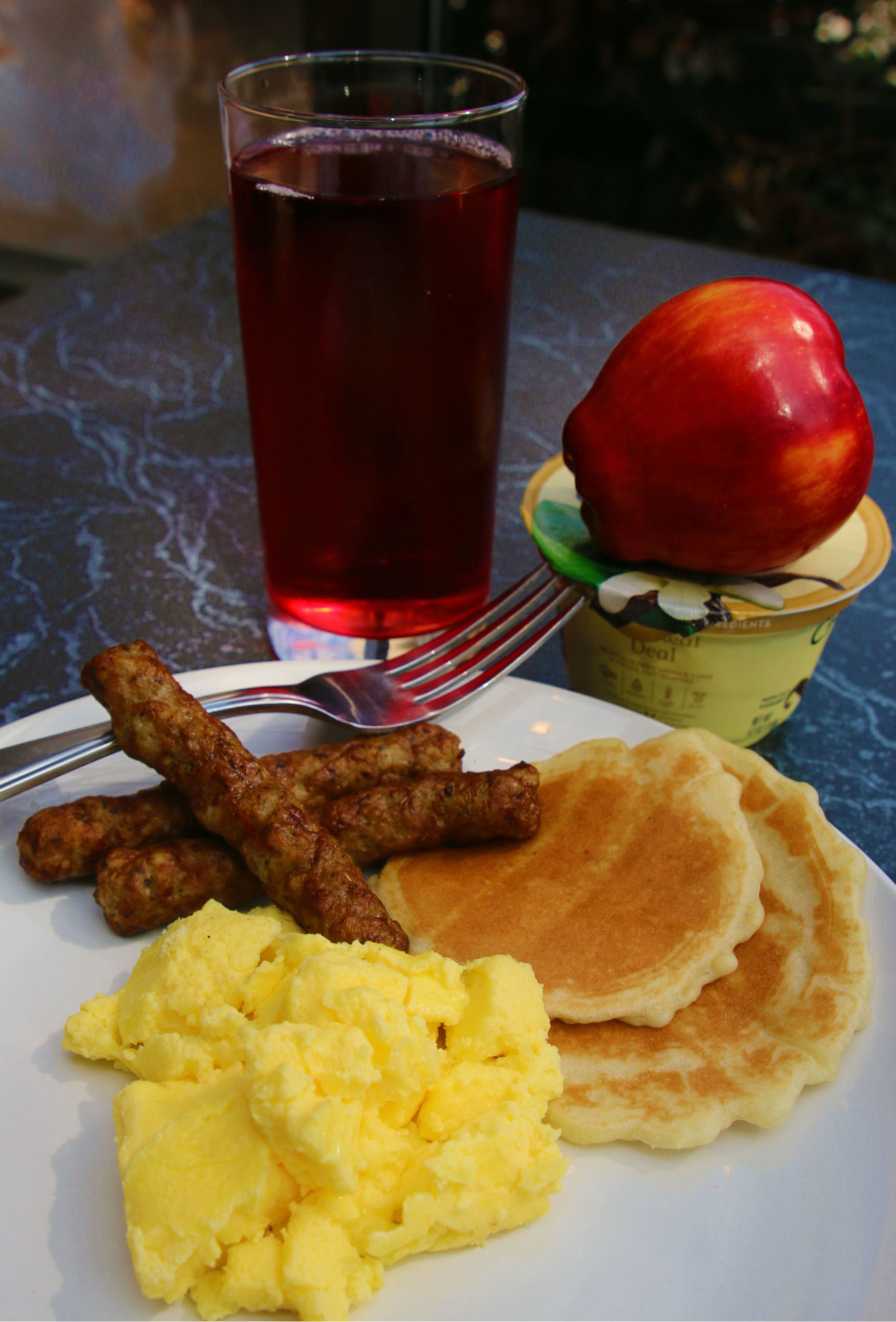 A plate with scrambled eggs, pancakes, sausages, and a glass of red beverage.