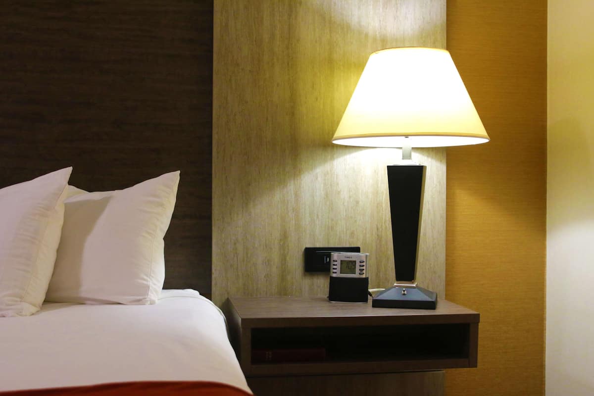 A neatly made bed with white linens and pillows, a bedside table with a lamp, a clock, and a small electronic device.