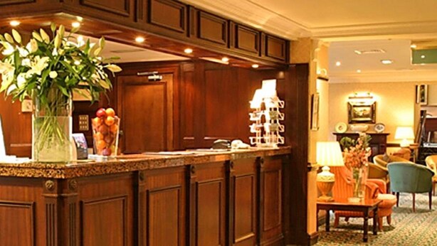 Un hall de hotel con mostrador de recepción, paneles de madera, una máquina de café y zona de estar con sillas y una mesa.