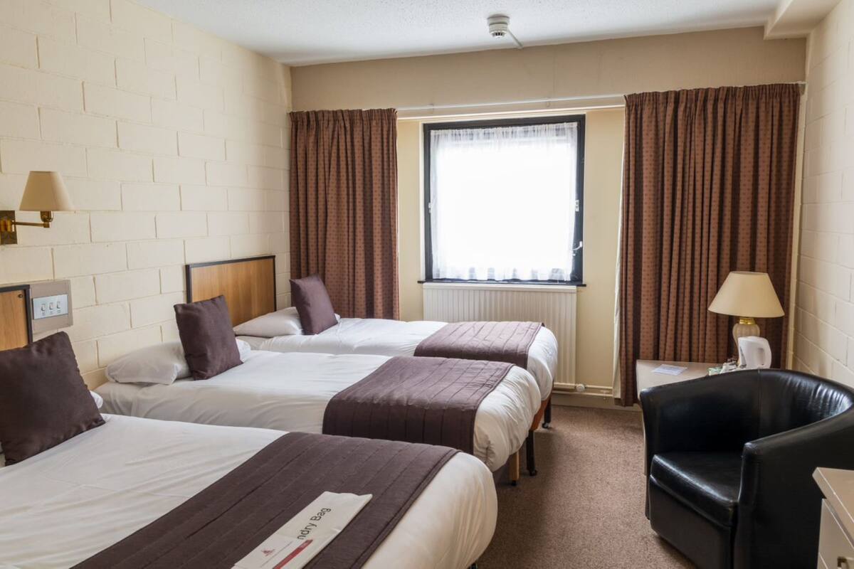 Hotellihuone, jossa on kolme erillistä sänkyä, musta nahkainen nojatuoli, pieni pöytä lampun kanssa ja ikkuna, jossa on verhot.