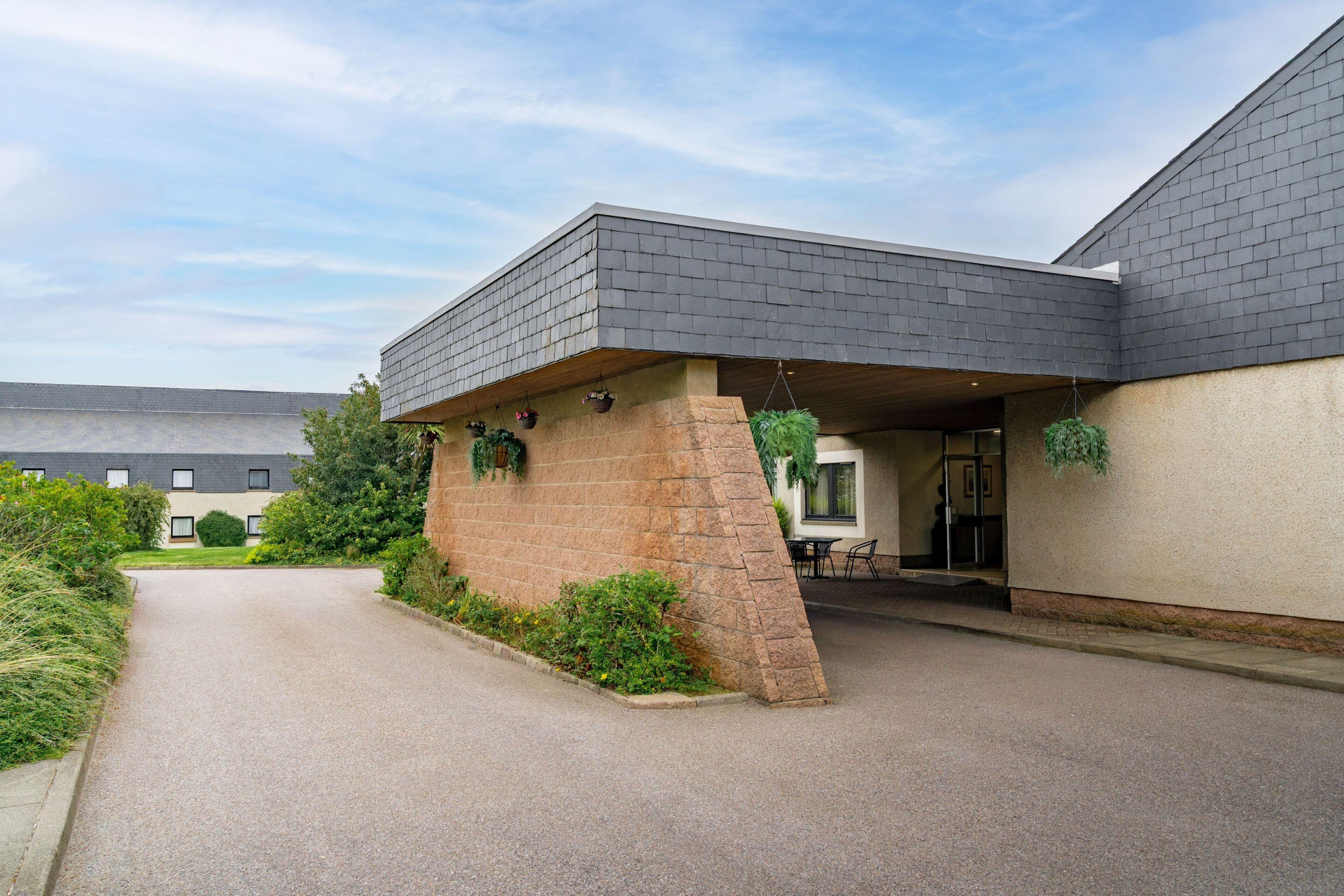A building with a flat roof, a covered entrance with a table and chairs, and a gravel driveway.