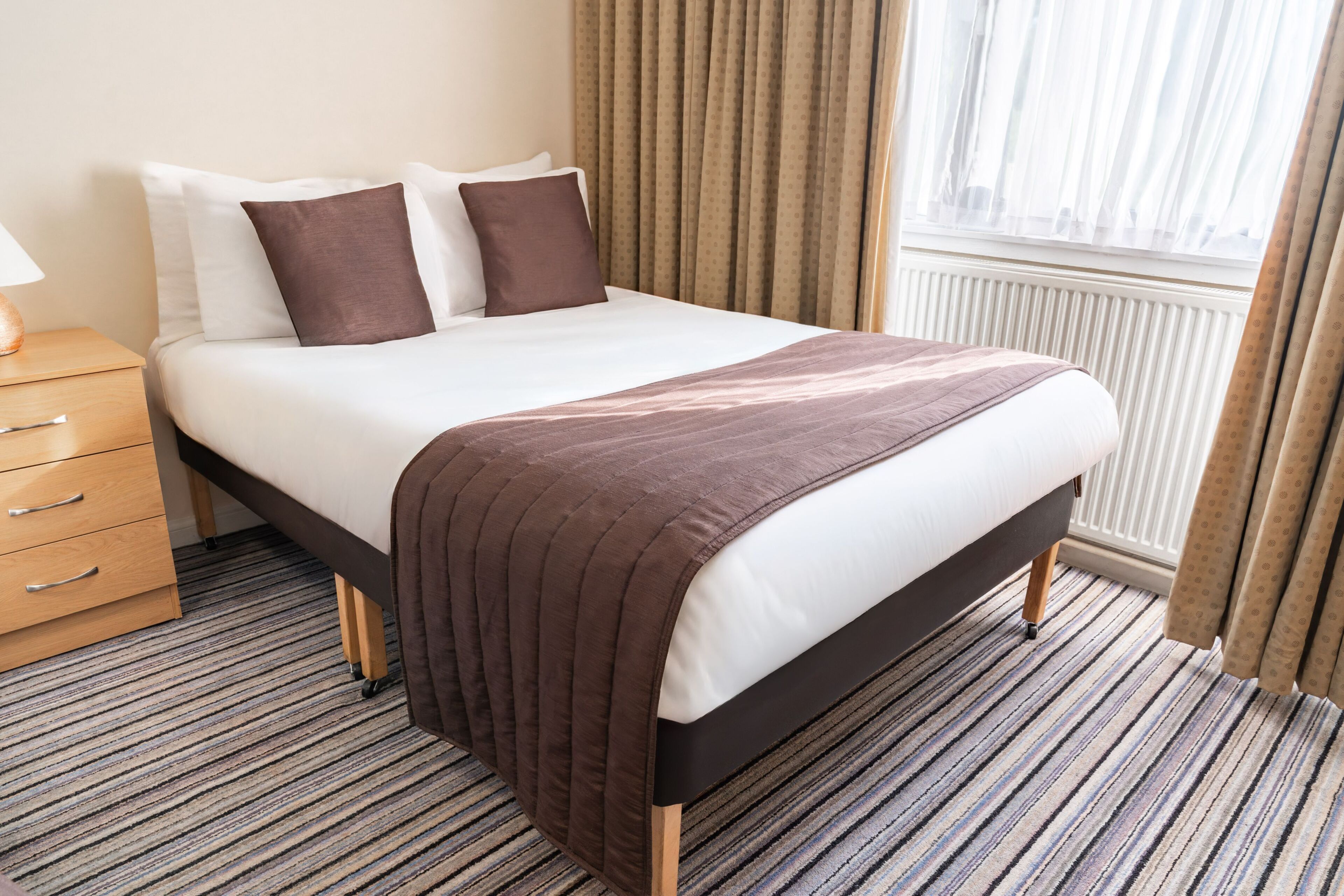 Une chambre d’hôtel avec un lit, une table de chevet, une lampe, une fenêtre avec des rideaux et un radiateur.