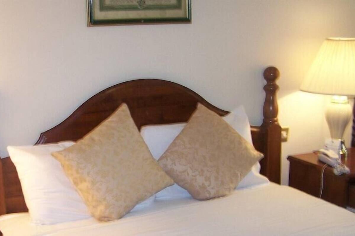 Hotellihuone, jossa on puinen sängynpääty, kaksi yöpöytävalaisinta, valkoisilla lakanoiden peitetty sänky ja seinällä oleva kehystetty kuva.