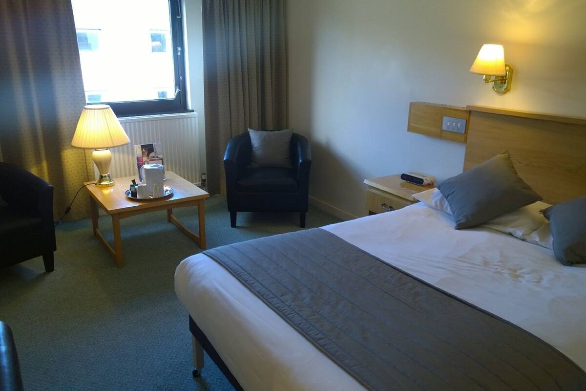 Hotellihuone, jossa on sänky, kirjoituspöytä lampun kanssa, tuoli ja yöpöytä.
