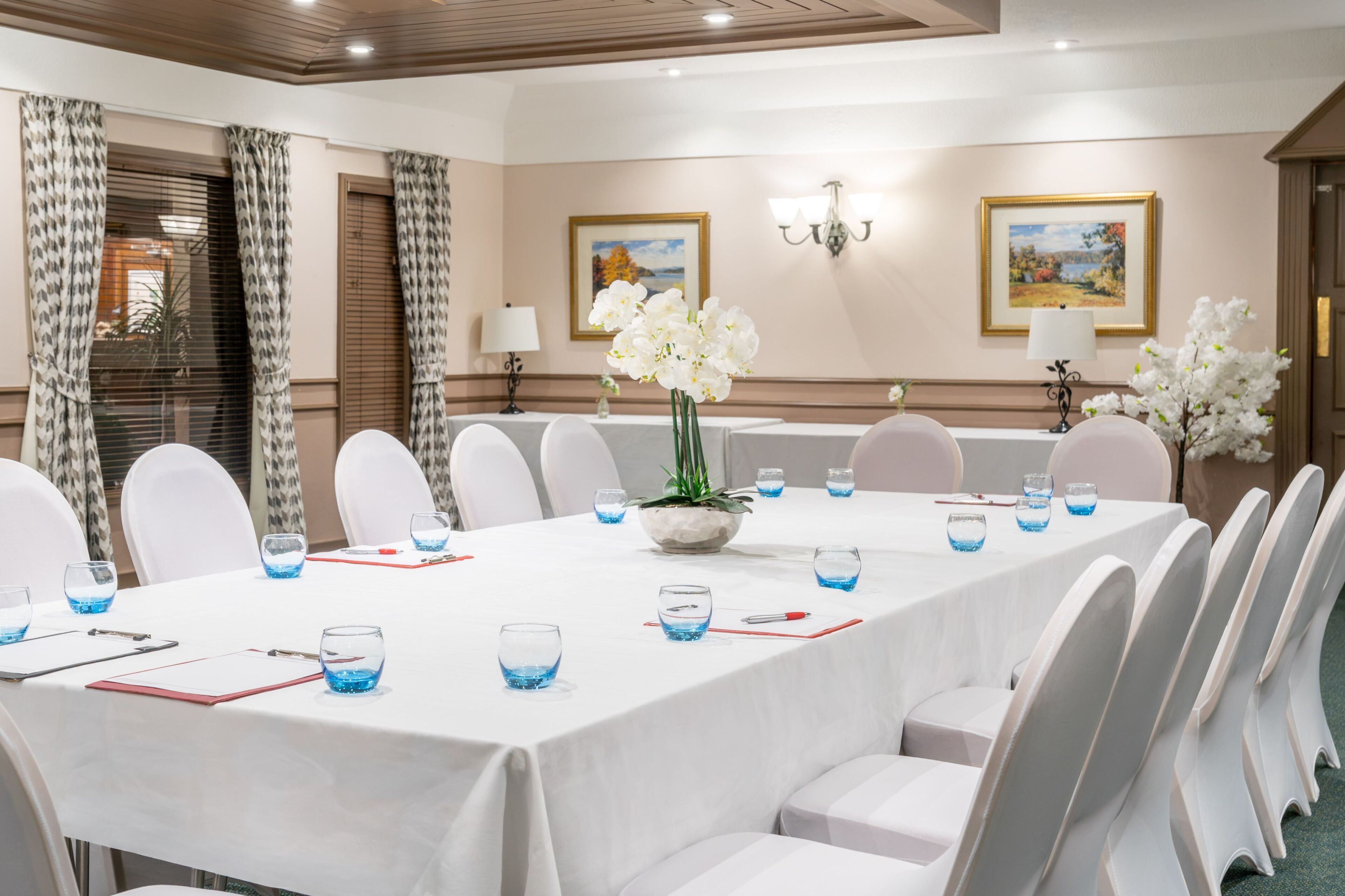 Een vergaderzaal met een lange tafel, witte stoelen, blauwe glazen waterbekers, een wit orchidee-bloemstuk en lijstwerken aan de wanden.