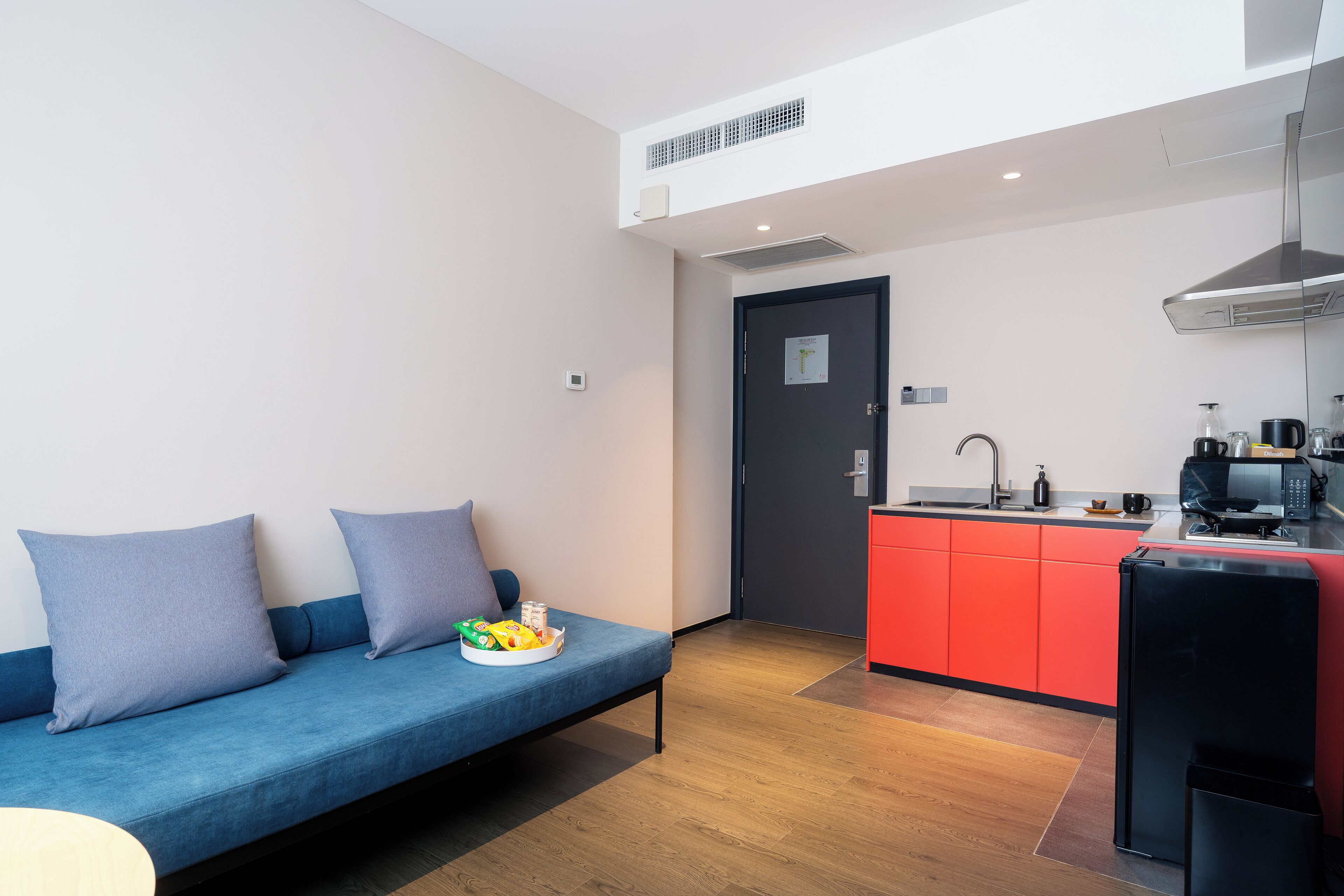 A modern living room with a blue sofa, a small kitchen area with red cabinets, and a black refrigerator.