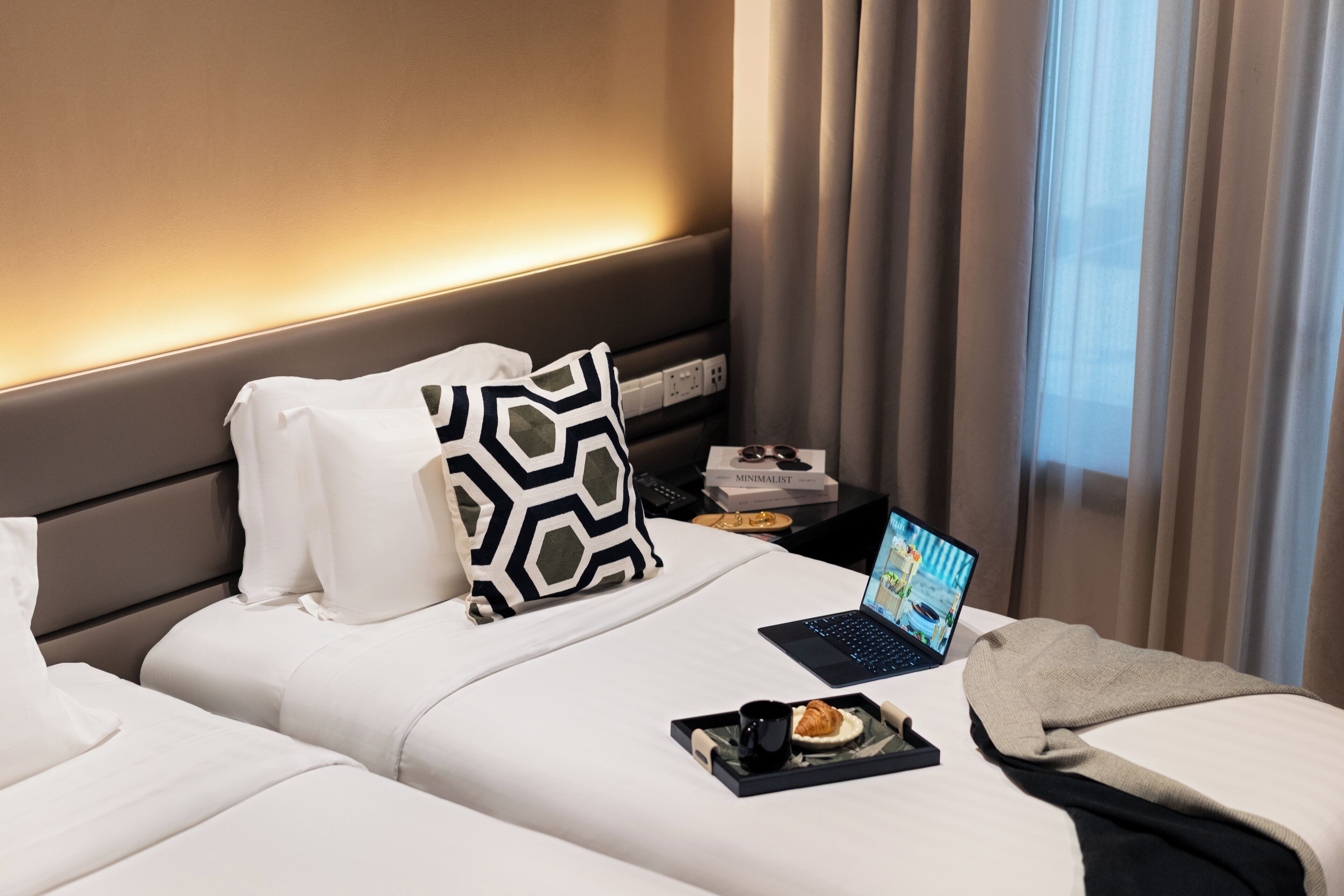 A hotel room with a bed, a laptop, a tray with food and a drink, and a book on a nightstand.