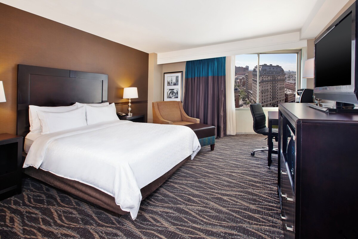 Quarto de hotel com uma cama grande, uma escrivaninha com uma TV, uma cadeira, uma luminária de mesa e vista para a cidade.