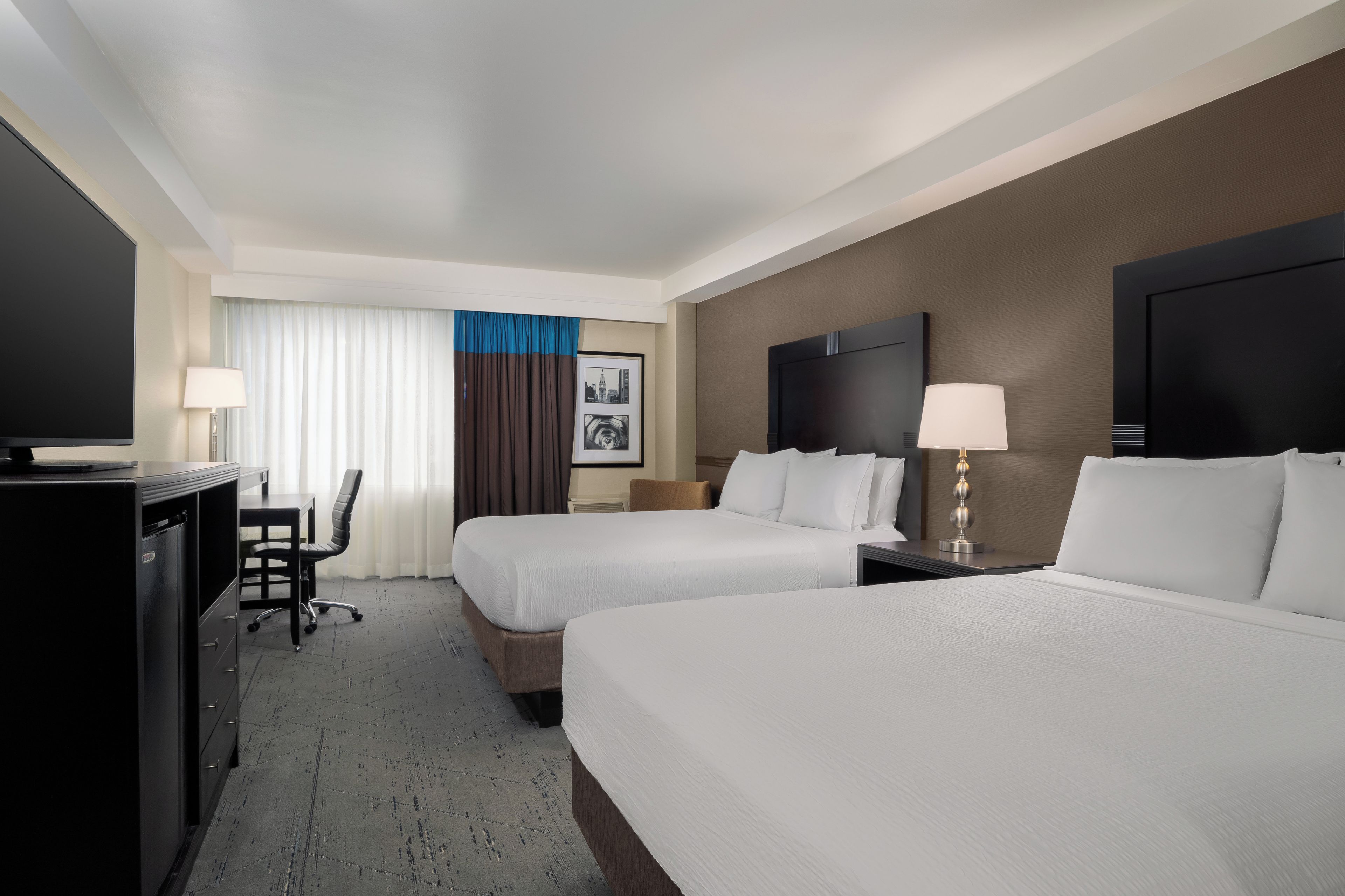 Ein Hotelzimmer mit zwei Betten, einem Schreibtisch, einem Sessel, einem Fernseher und einem gerahmten Bild an der Wand.