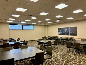 Sala de conferência com várias mesas e cadeiras, carpete estampado, luminárias no teto e uma pintura da paisagem urbana na parede.