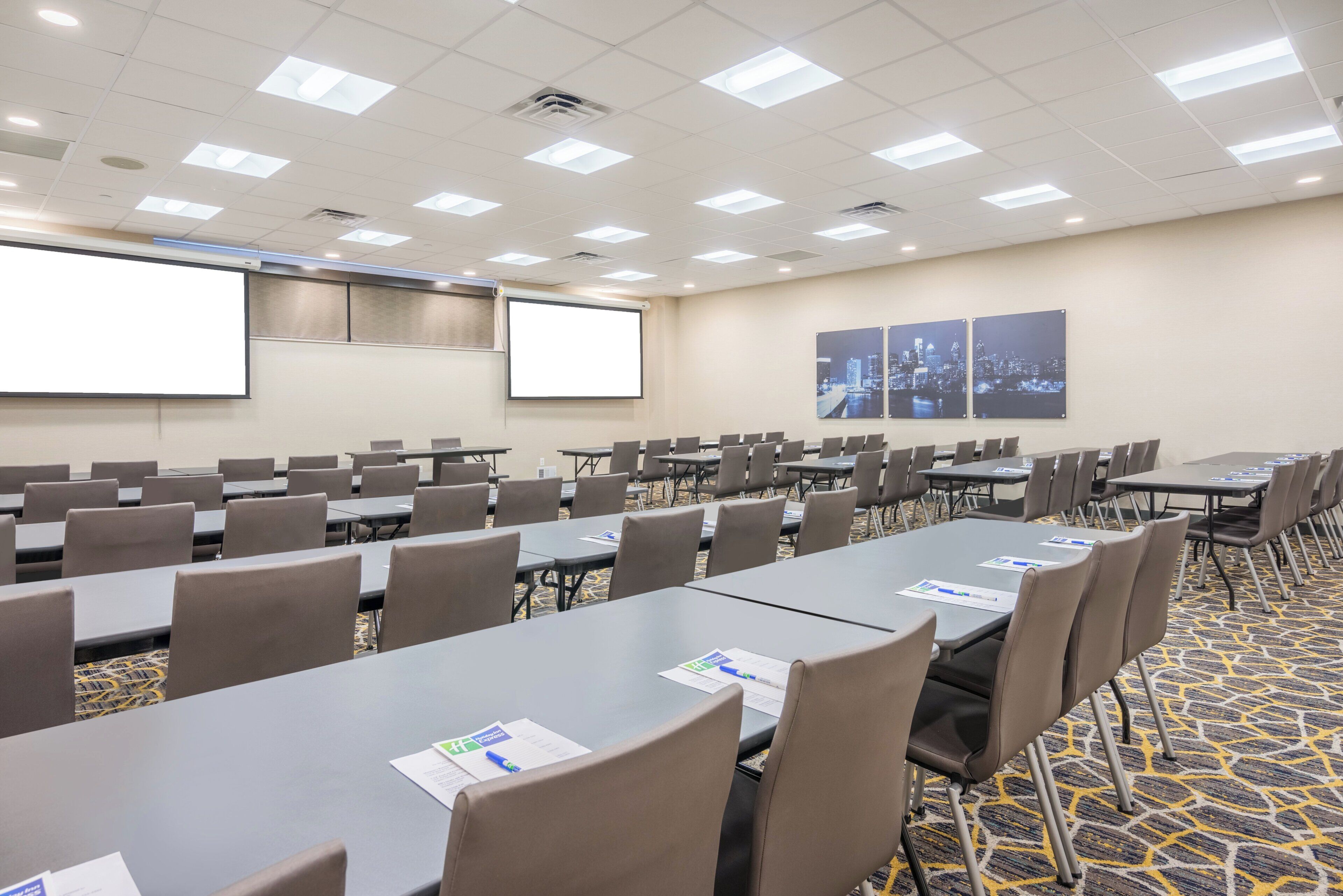 Un salón de conferencias con varias mesas y sillas, una pantalla de proyector y dos pinturas de paisajes urbanos en la pared.