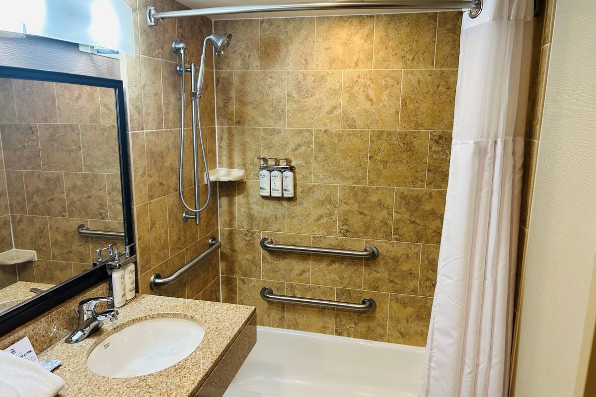 Un bagno con lavandino, specchio, doccia e maniglie di sostegno.