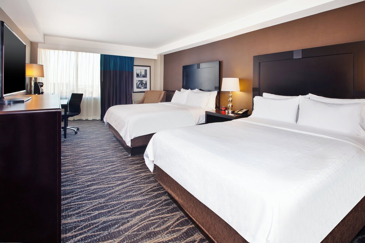 Quarto de hotel com duas camas, uma escrivaninha, uma cadeira, televisão e piso coberto por carpete.