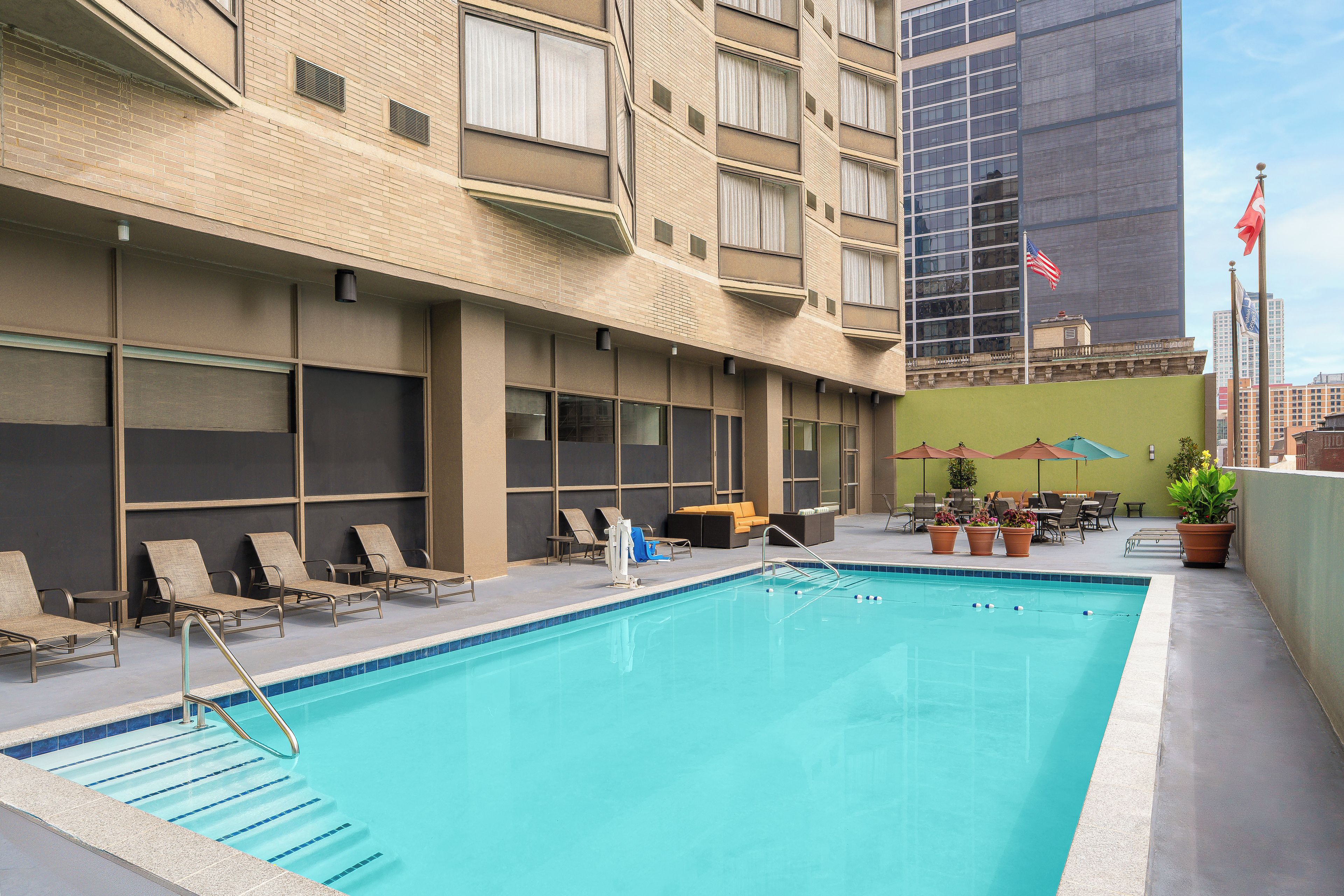 A swimming pool with lounge chairs and umbrellas, surrounded by a building with multiple windows and a flag.