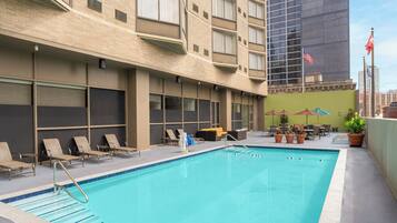 A swimming pool with lounge chairs and umbrellas, surrounded by a building with multiple windows and a flag.