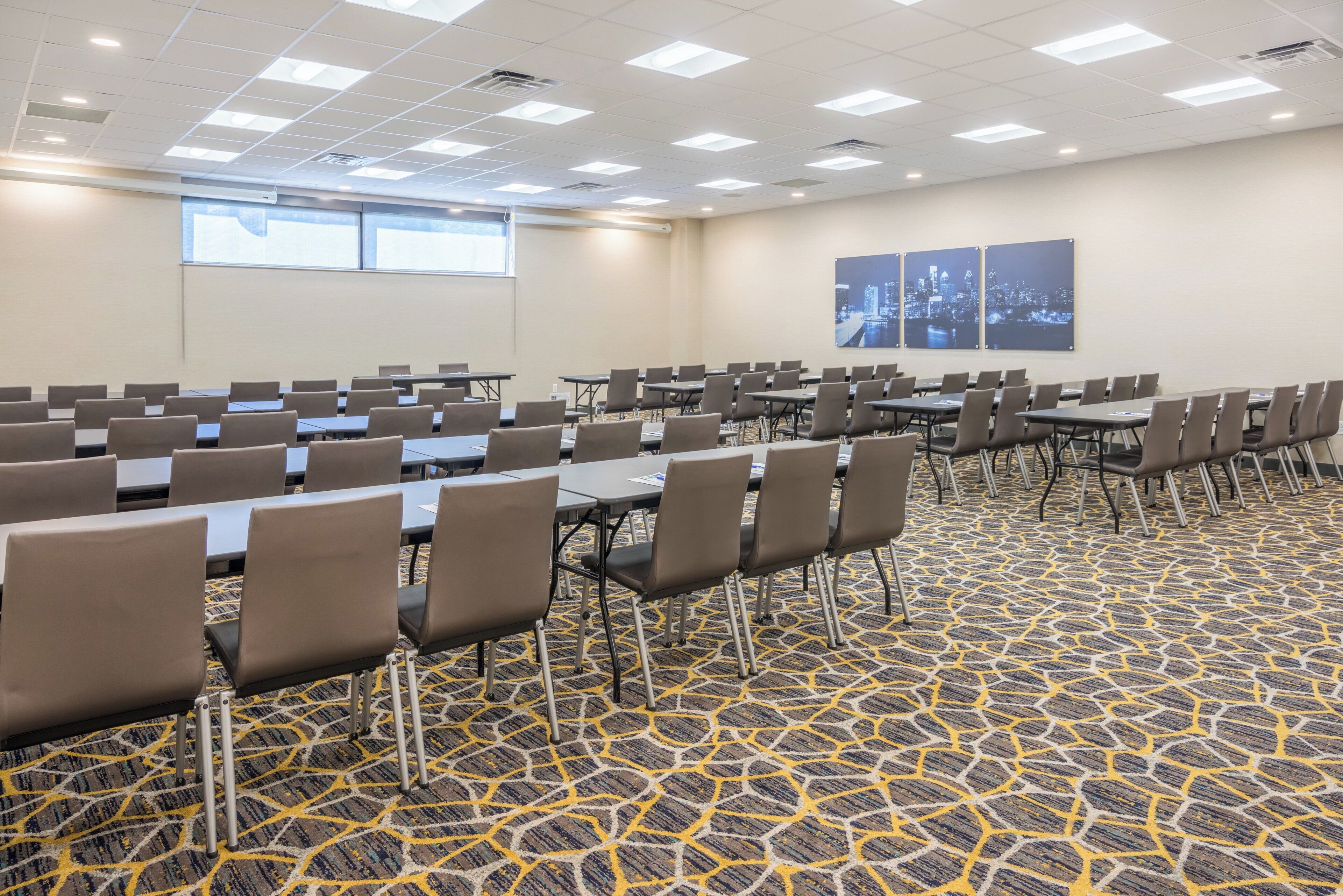 A conference room with rows of tables and chairs, carpeted floor, and a cityscape view.