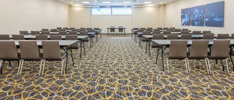 Une salle de conférence avec un tapis à motifs, des rangées de tables et de chaises, une grande fenêtre et une affiche représentant un paysage urbain accrochée au mur.