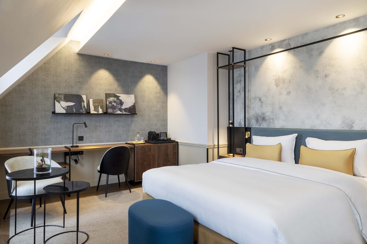 A modern hotel room with a large bed, a desk with a chair, a small table with a glass container, and artwork on the wall.