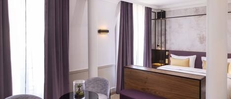 A hotel room with a bed, a bedside table, a chair, a small round table, and a bench.