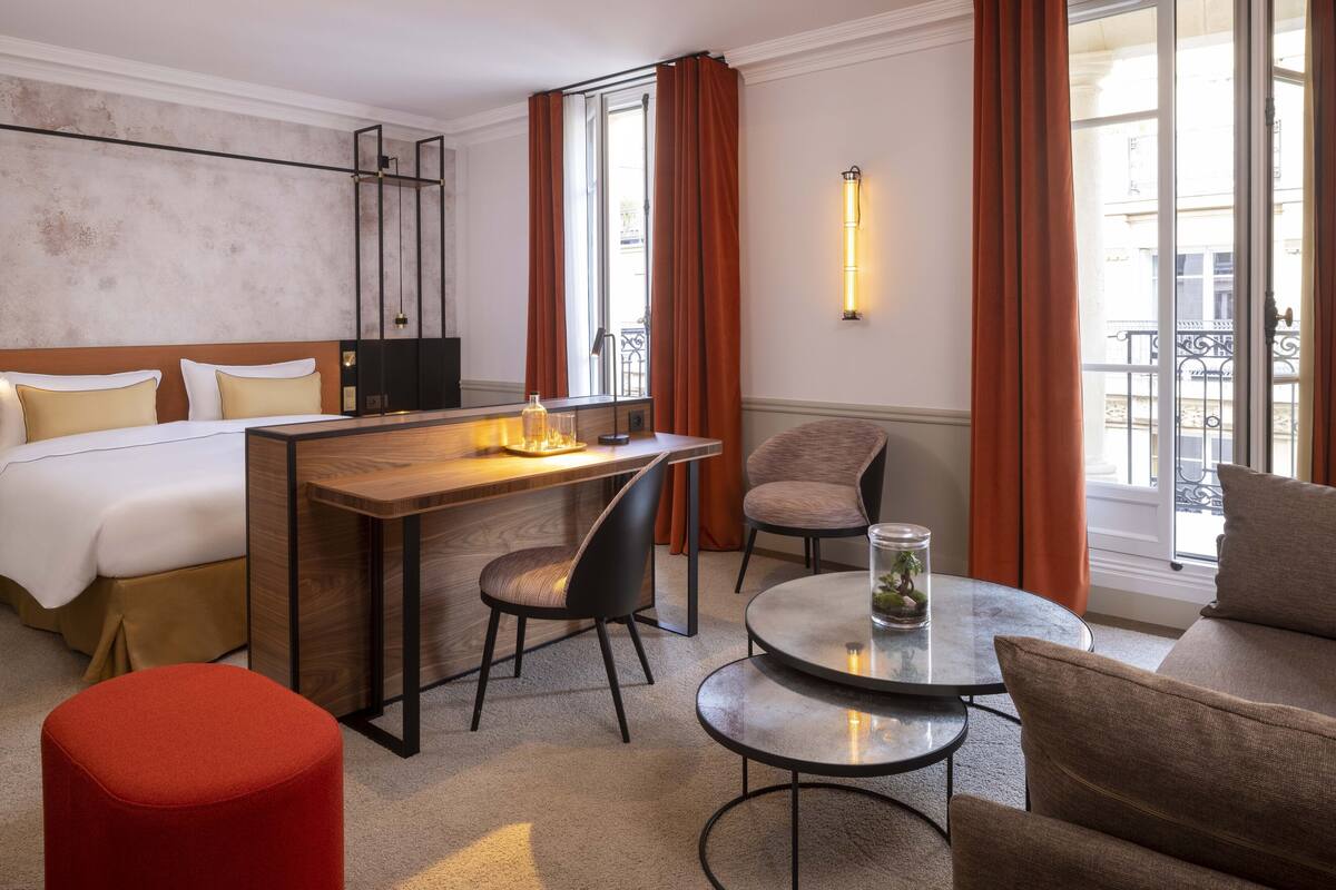 A hotel room with a bed, a desk, a chair, a round table, and a red ottoman.
