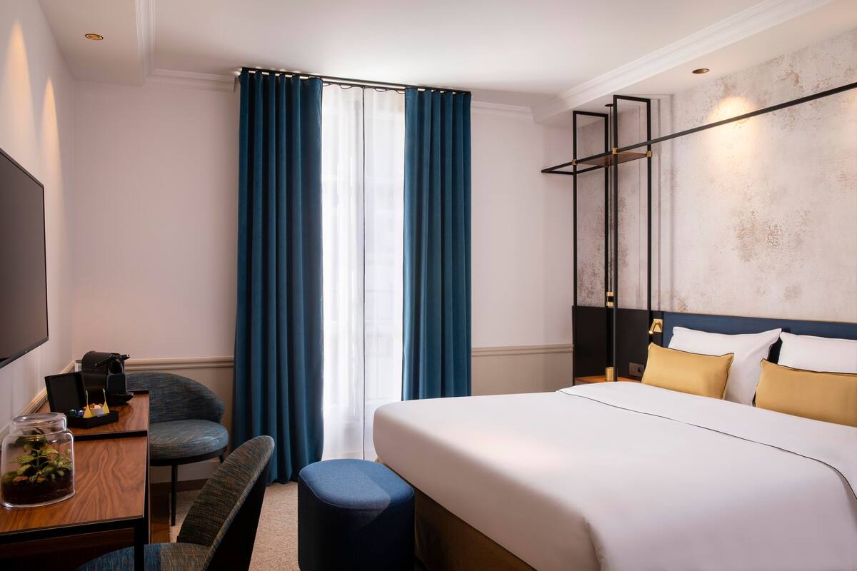 A hotel room with a bed, a desk with a chair, a TV, and a window with curtains.