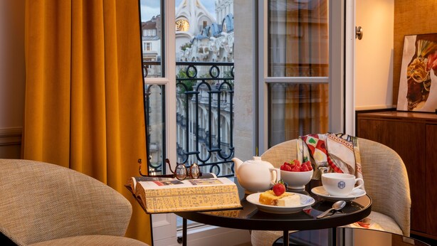 A room with a table set for breakfast, a view of a building with a clock tower, and a balcony with a wrought iron railing.