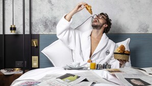 A hotel room with a bed, a bedside table with a newspaper, a basket of croissants, a cup of coffee, and a glass of orange juice.
