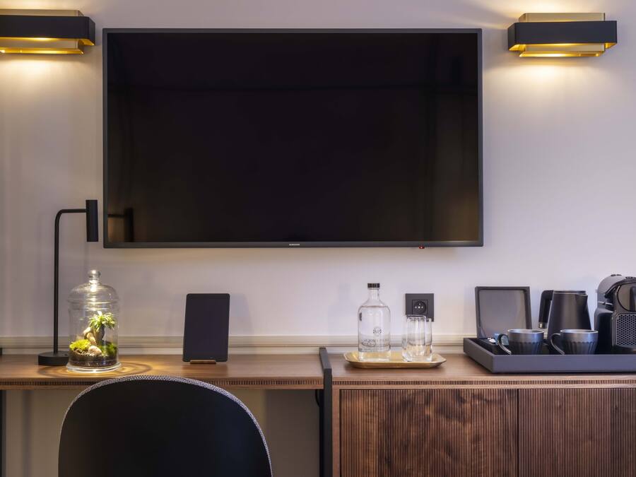 Une chambre d’hôtel moderne équipée d’un grand téléviseur à écran plat fixé au mur, d’un bureau en bois surmonté d’un terrarium en verre, d’un haut-parleur, d’une machine à café et d’un plateau avec des verres et une bouteille.