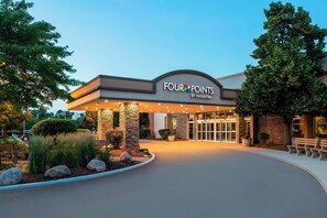 L’entrée d’un hôtel, avec une enseigne indiquant « FOUR POINTS BY SHERATON », présente un design moderne avec des piliers en pierre et un toit incurvé.