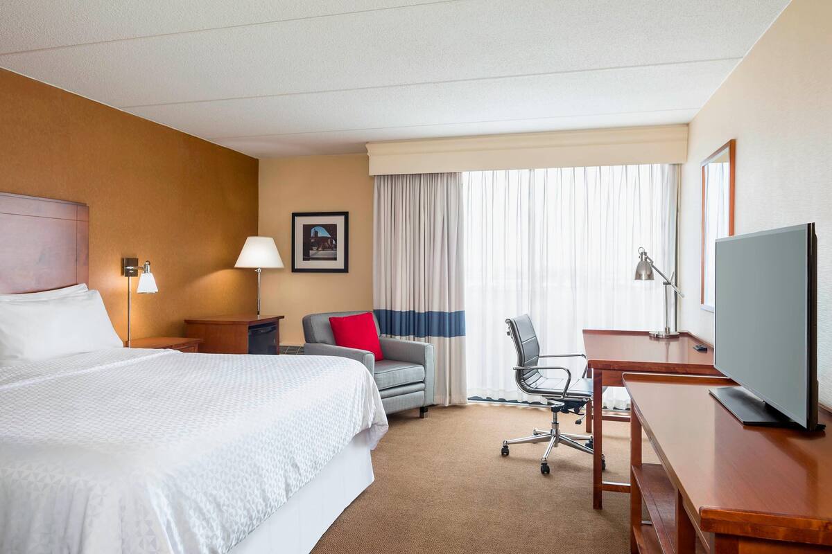 A hotel room with a large bed, a desk with a television, a chair, and a desk lamp.