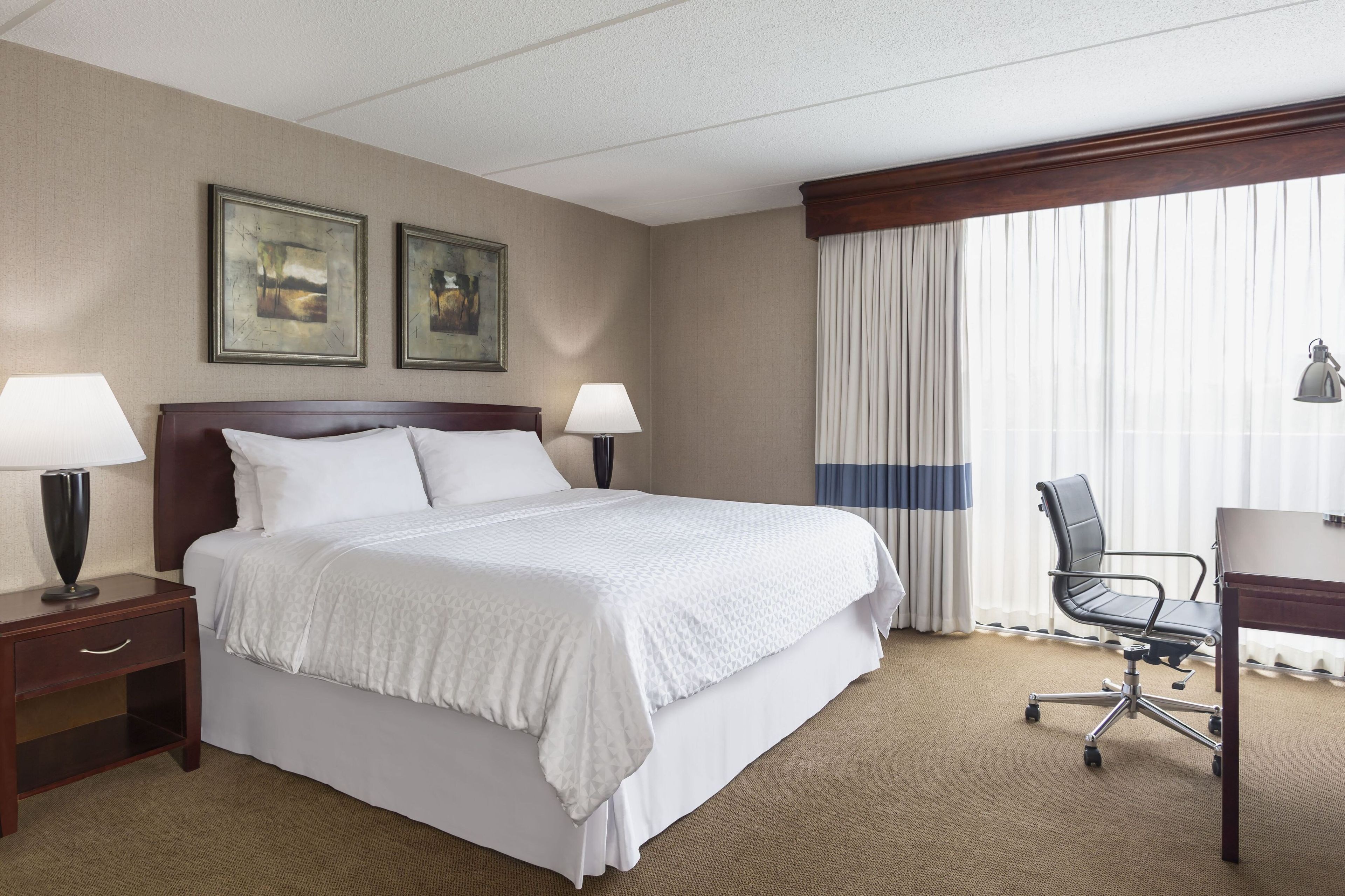 A hotel room with a large bed, two bedside tables with lamps, a desk with a chair, and a window with curtains.