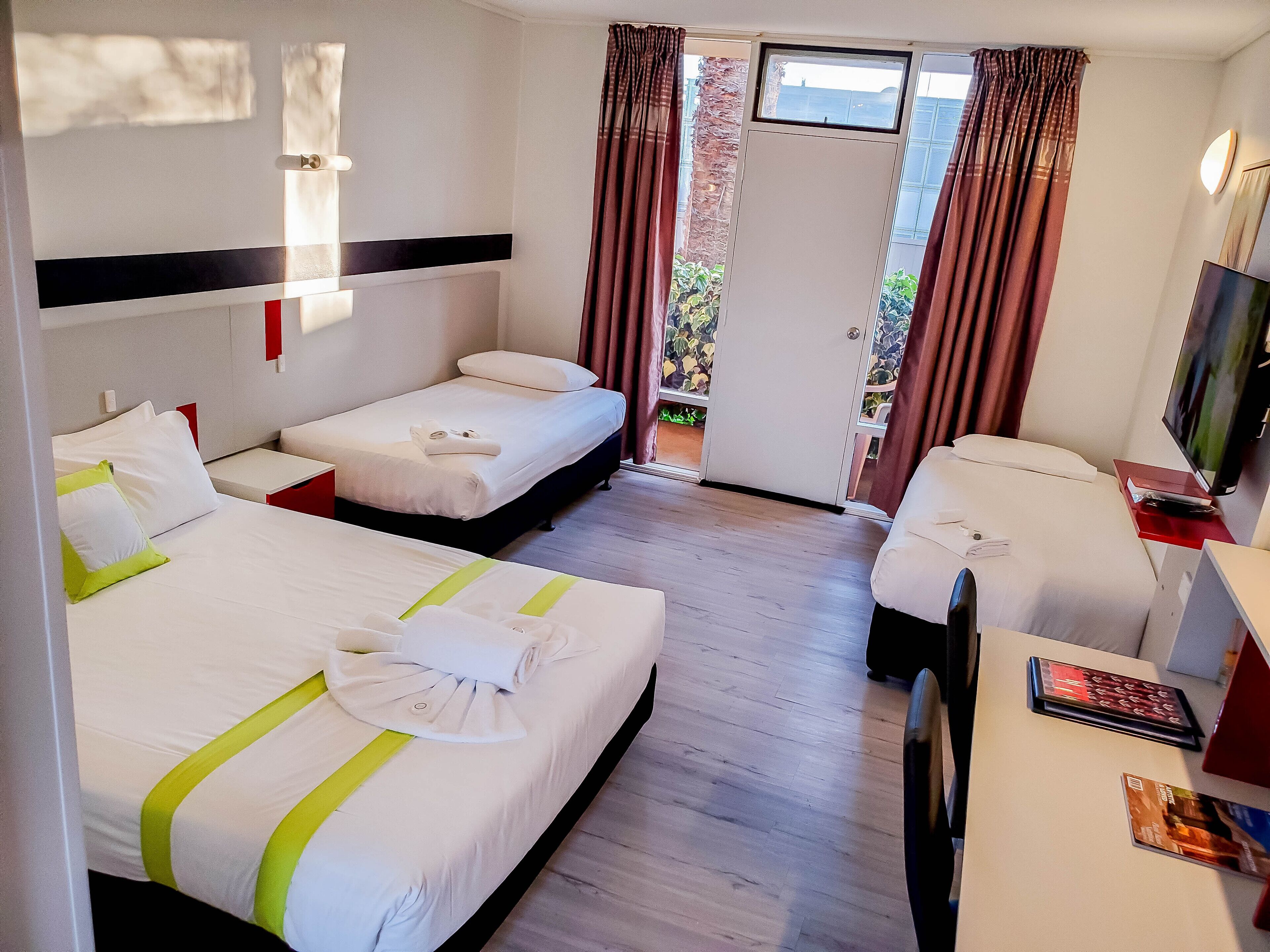 A hotel room with two beds, a desk, a TV, and a balcony with curtains.