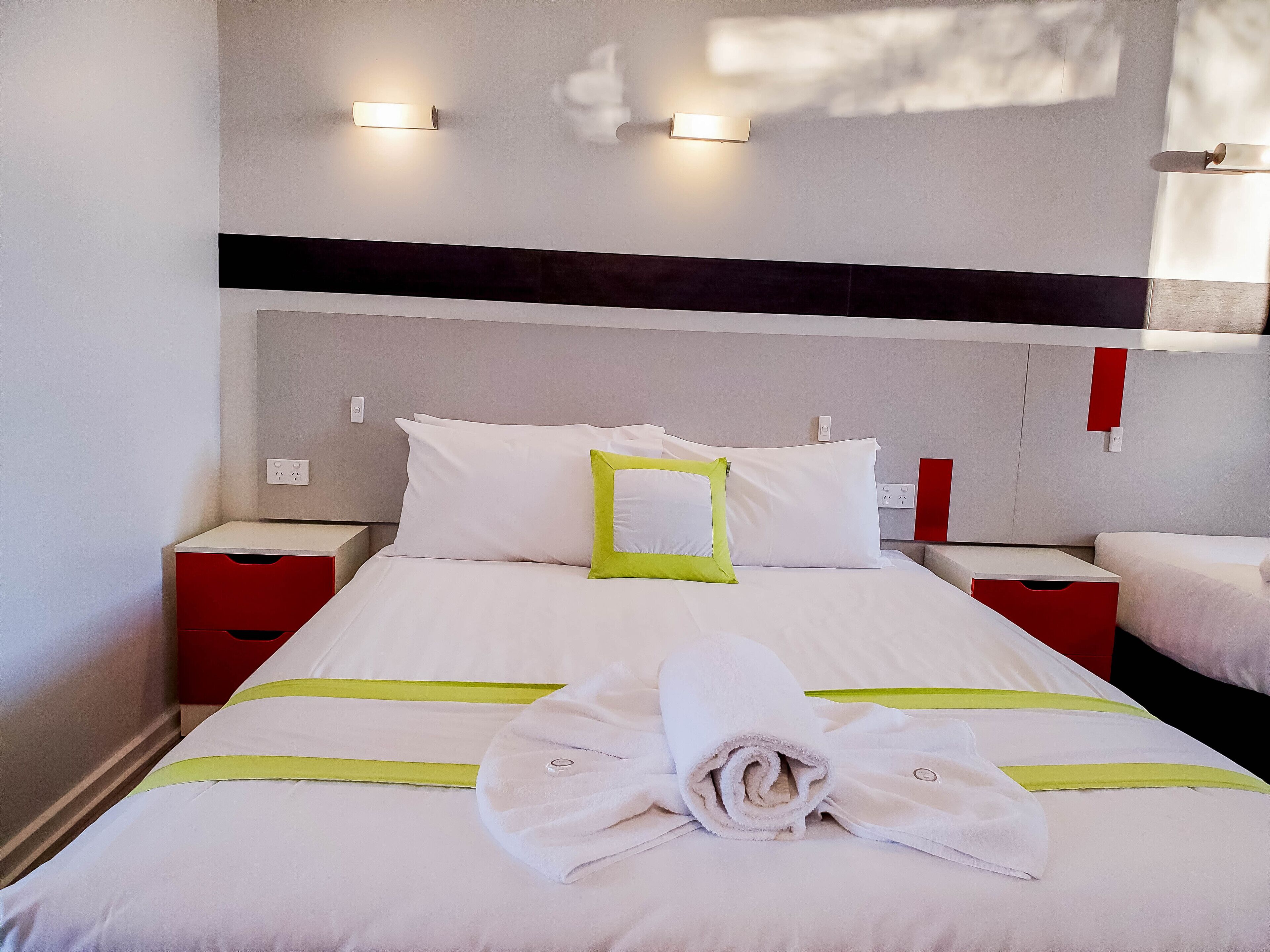 Una cama bien hecha con ropa de cama blanca y verde, dos mesitas de noche e iluminación empotrada en la pared.