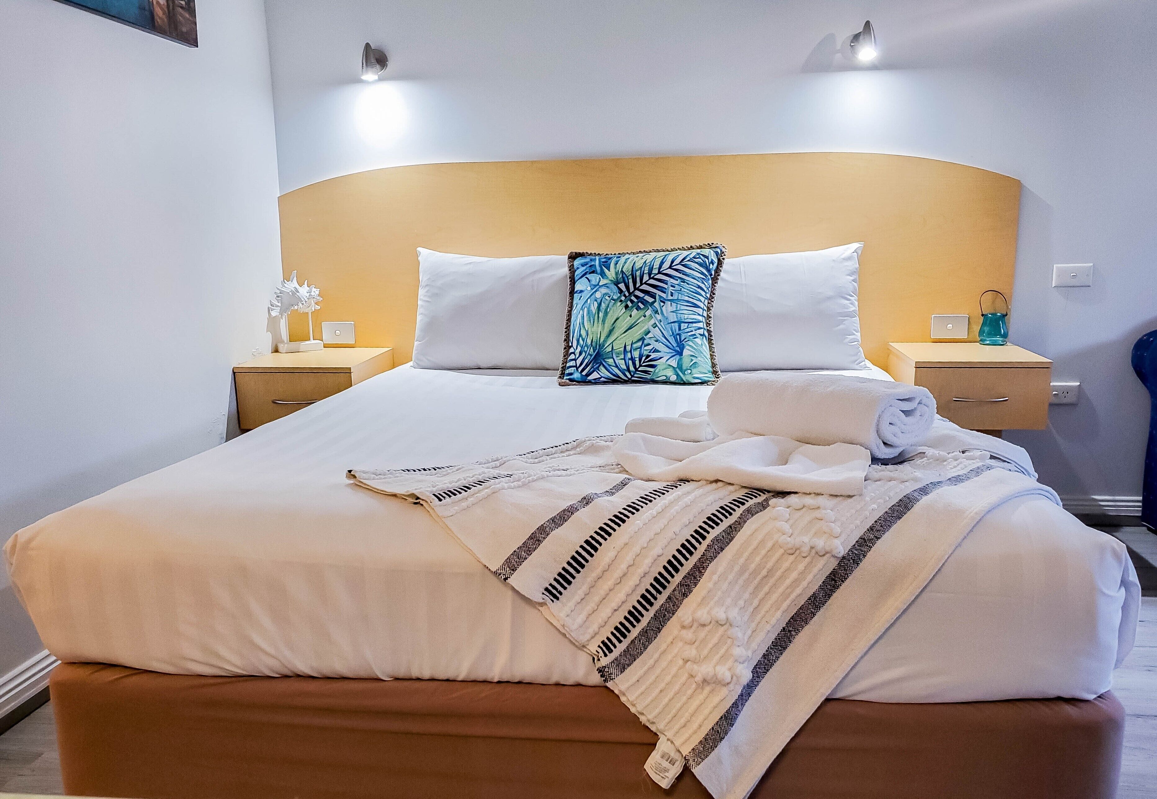 Un lit bien fait, avec du linge de lit blanc, un coussin orné d’une feuille tropicale bleue et un plaid rayé. Il y a deux tables de chevet avec des lampes, et un petit objet décoratif est posé sur l’une d’elles.