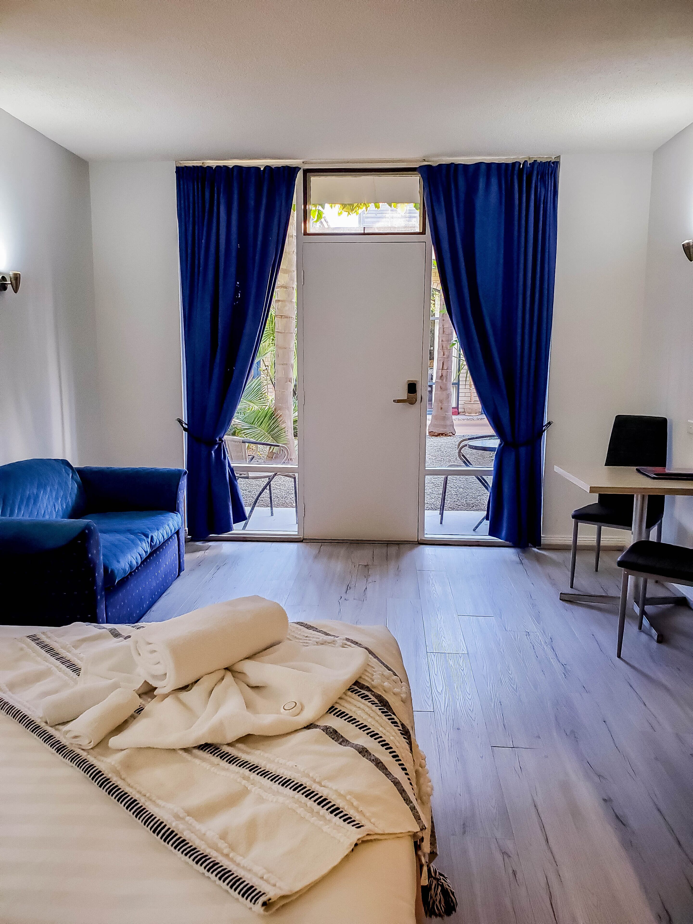A hotel room with a bed, a blue sofa, a desk with a chair, and a view through a door with blue curtains.