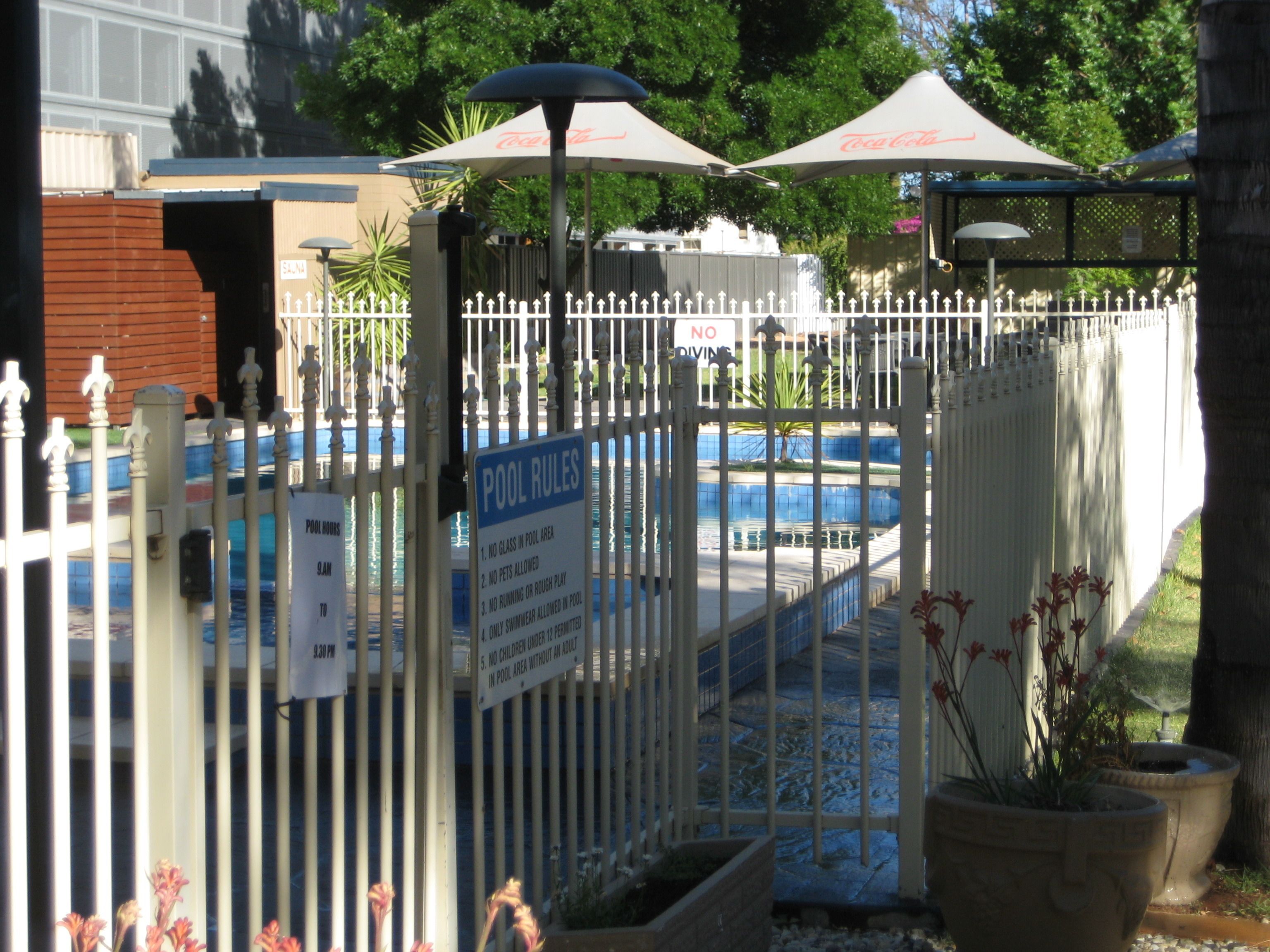 Zona de piscina amurallada con carteles de normas de la piscina, una sombrilla con el logo de Coca-Cola y plantas en macetas.