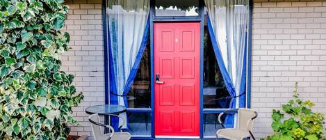 A red door with two windows, a white brick wall, and two wicker chairs outside.