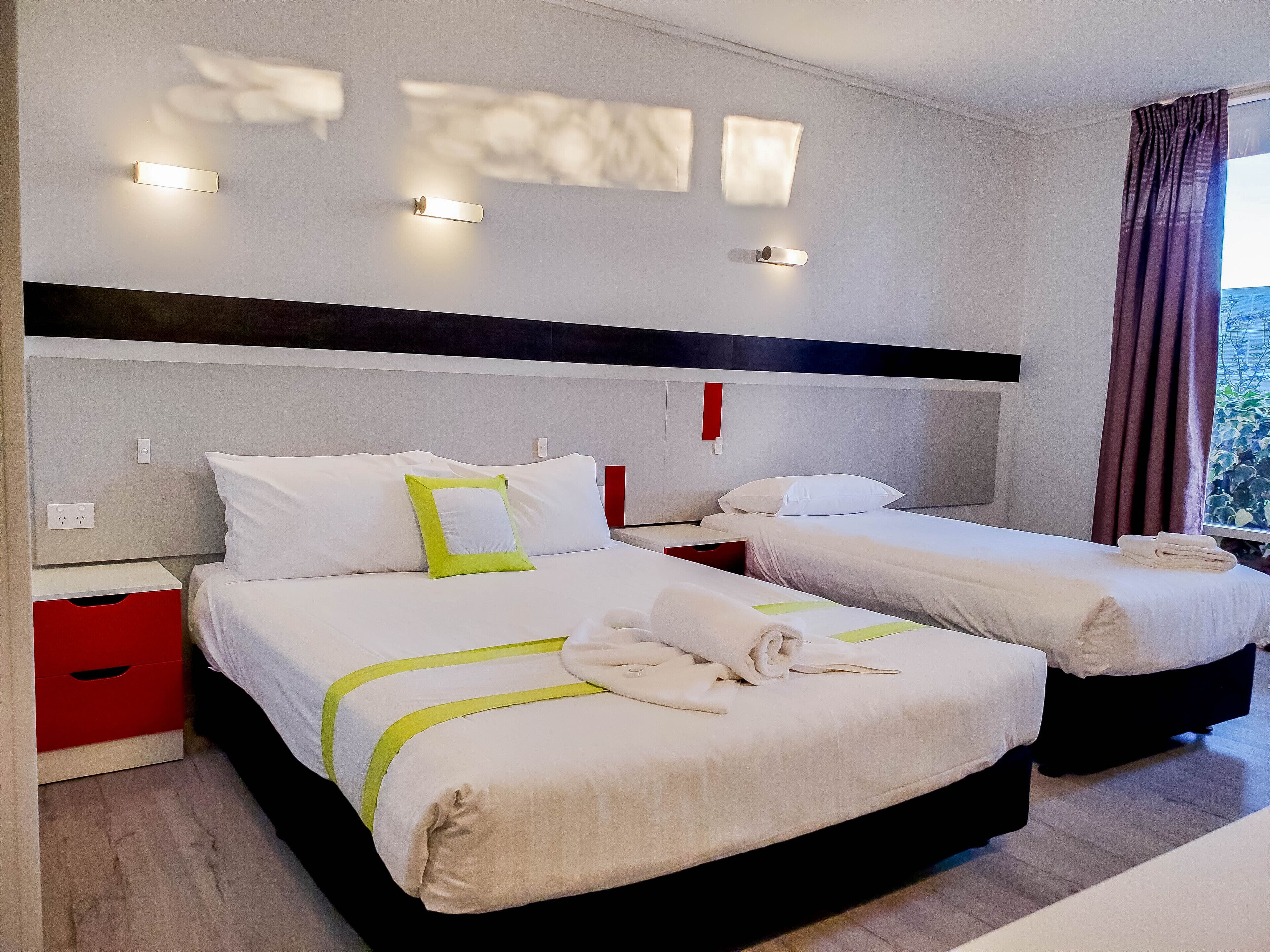 Een hotelkamer met twee bedden, wit beddengoed, een rode nachtkastje en uitzicht op groen buiten.